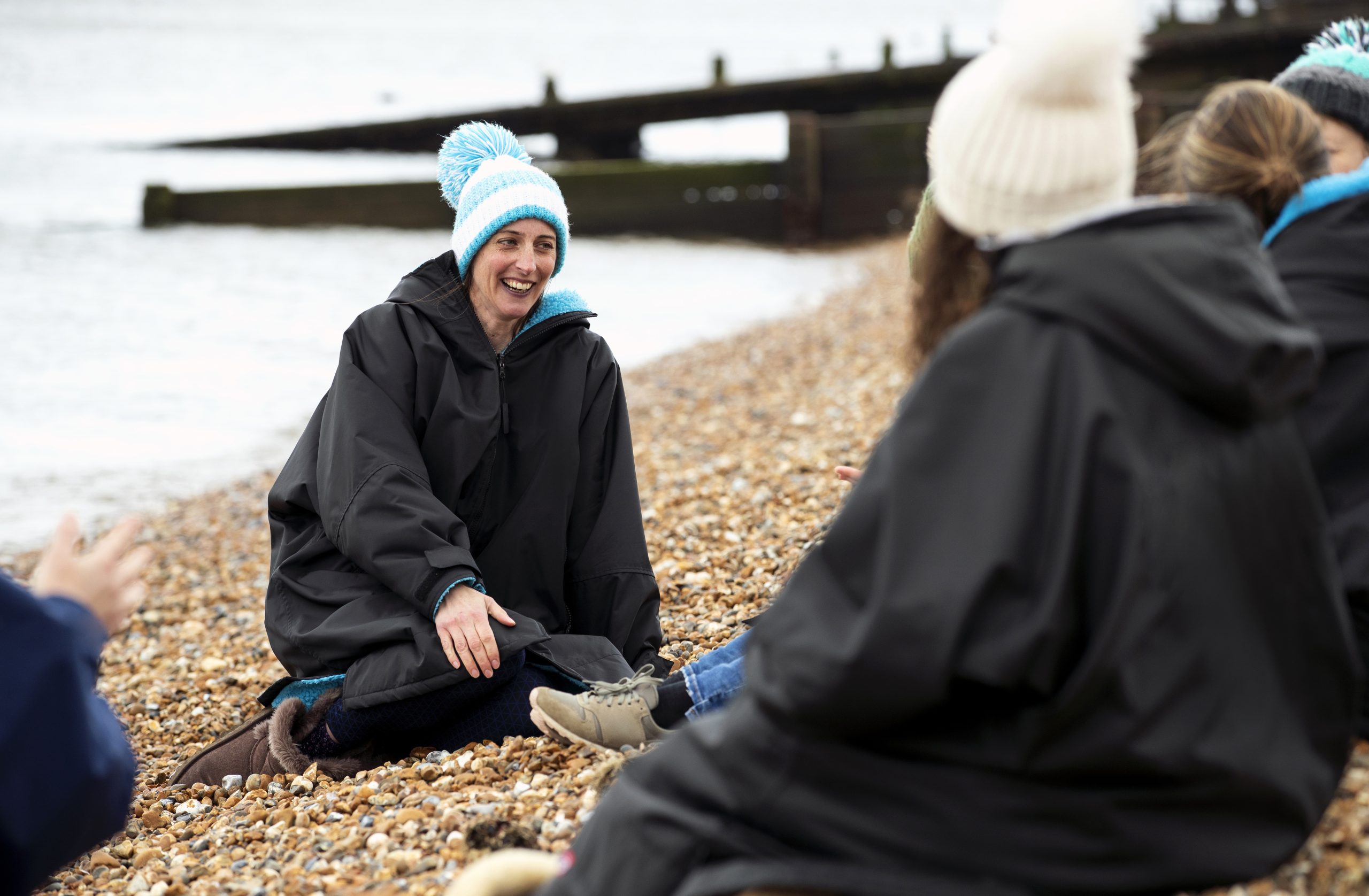 A group of women aged 40-60 sitting on a beach wrapped up after cold water swimming