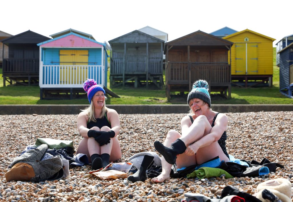 women in swimsuits and bobble hats sitting on a shingle beach in front of beach huts