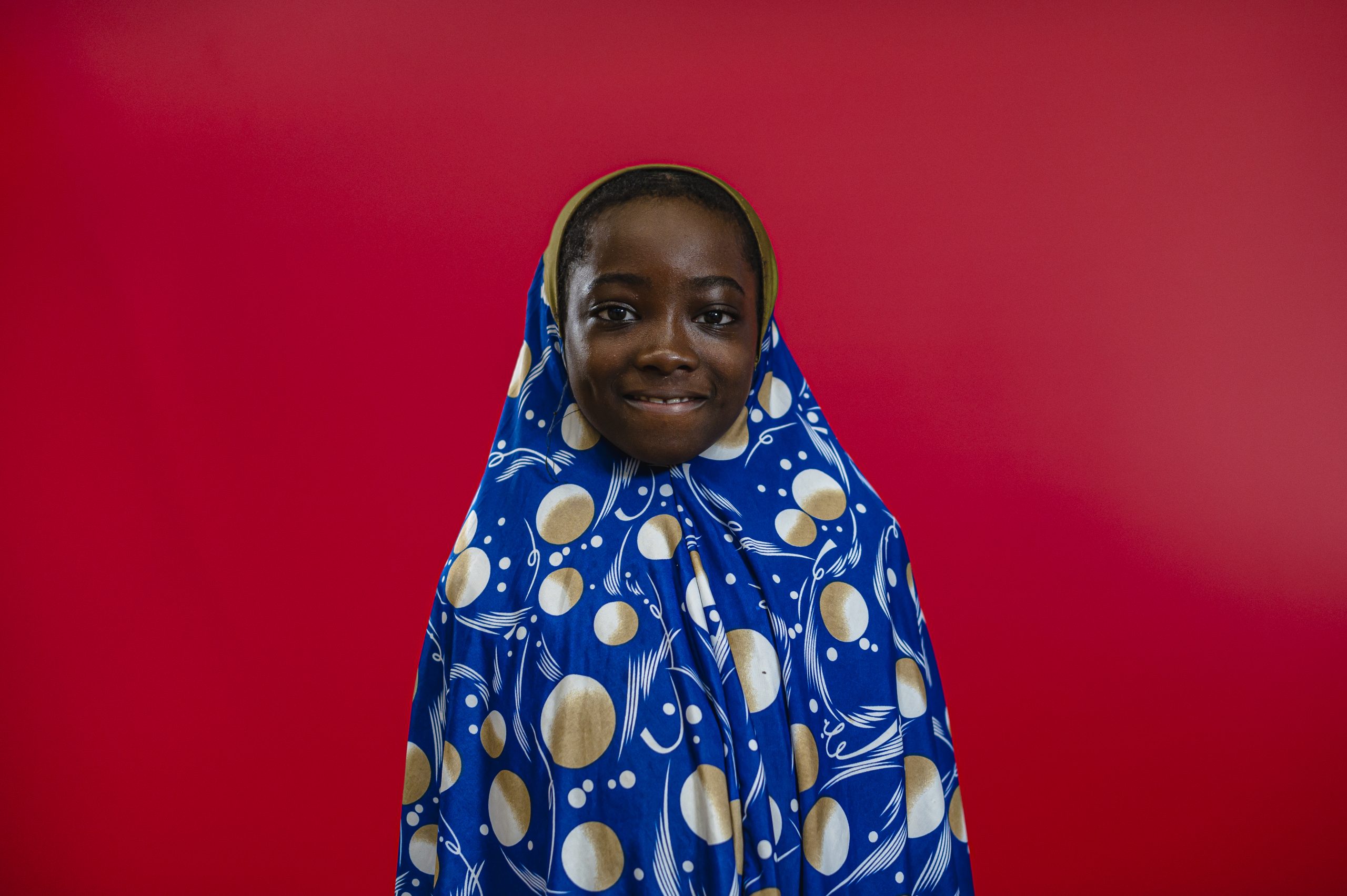 black primary school girl in blue hijab decorated with planets and stars. She is looking at the camera stood against a red background
