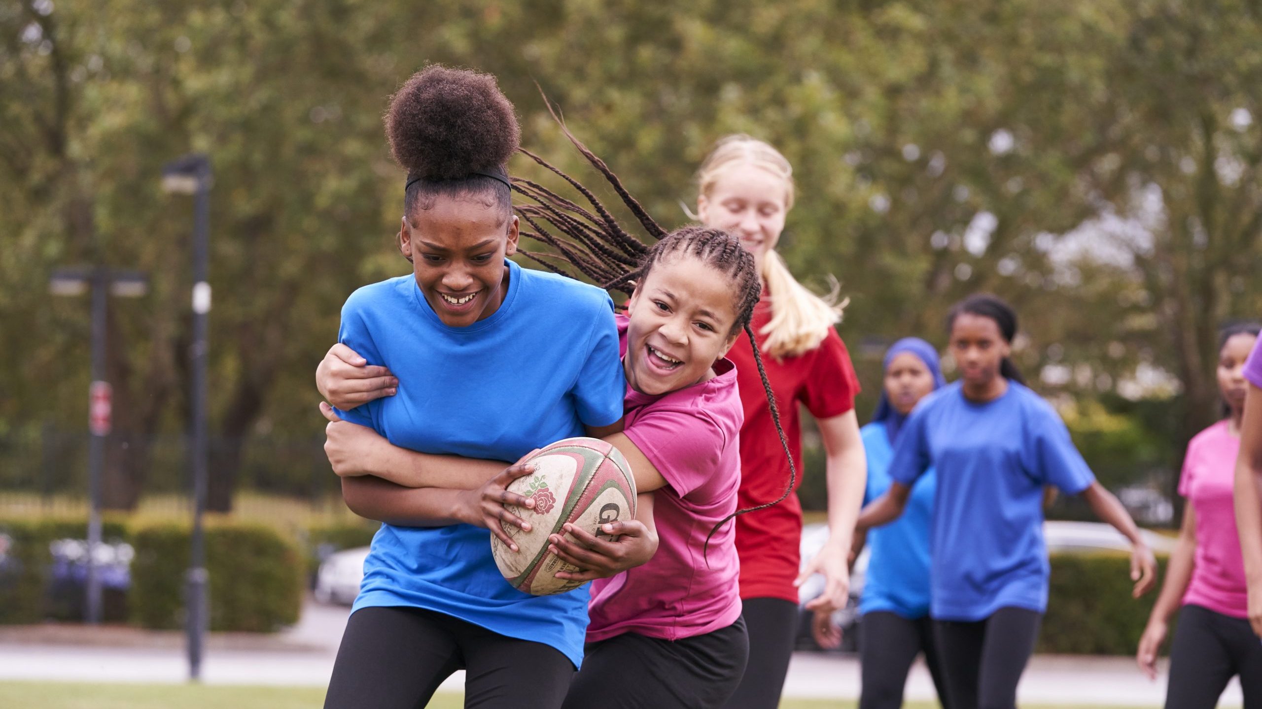 A group of high school girls playing rugby, two black girls tackling with smiles on their faces