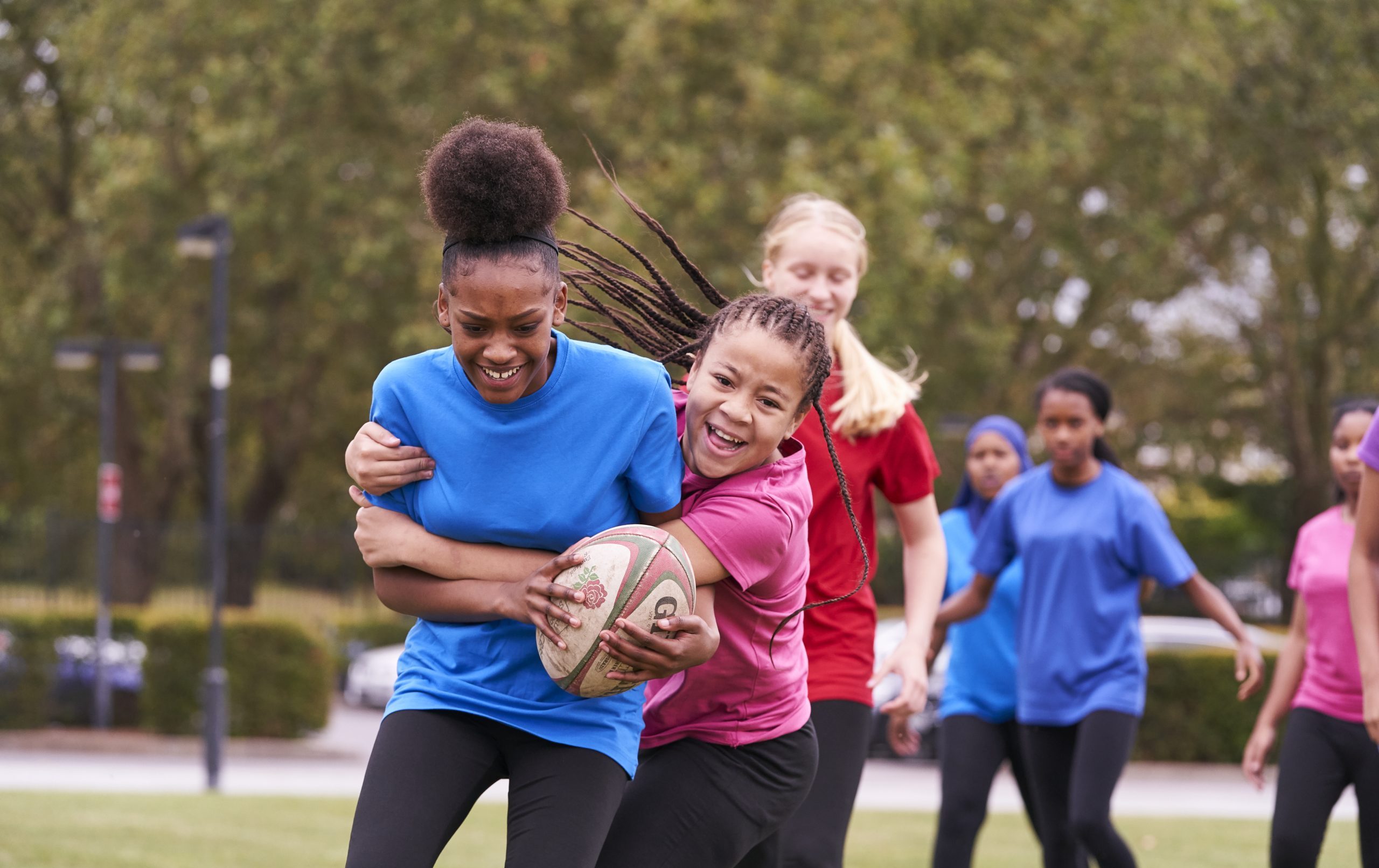 A group of high school girls playing rugby, two black girls tackling with smiles on their faces