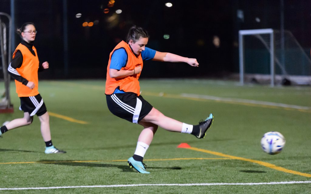 white girl in an orange bib kicking a football at a goal 