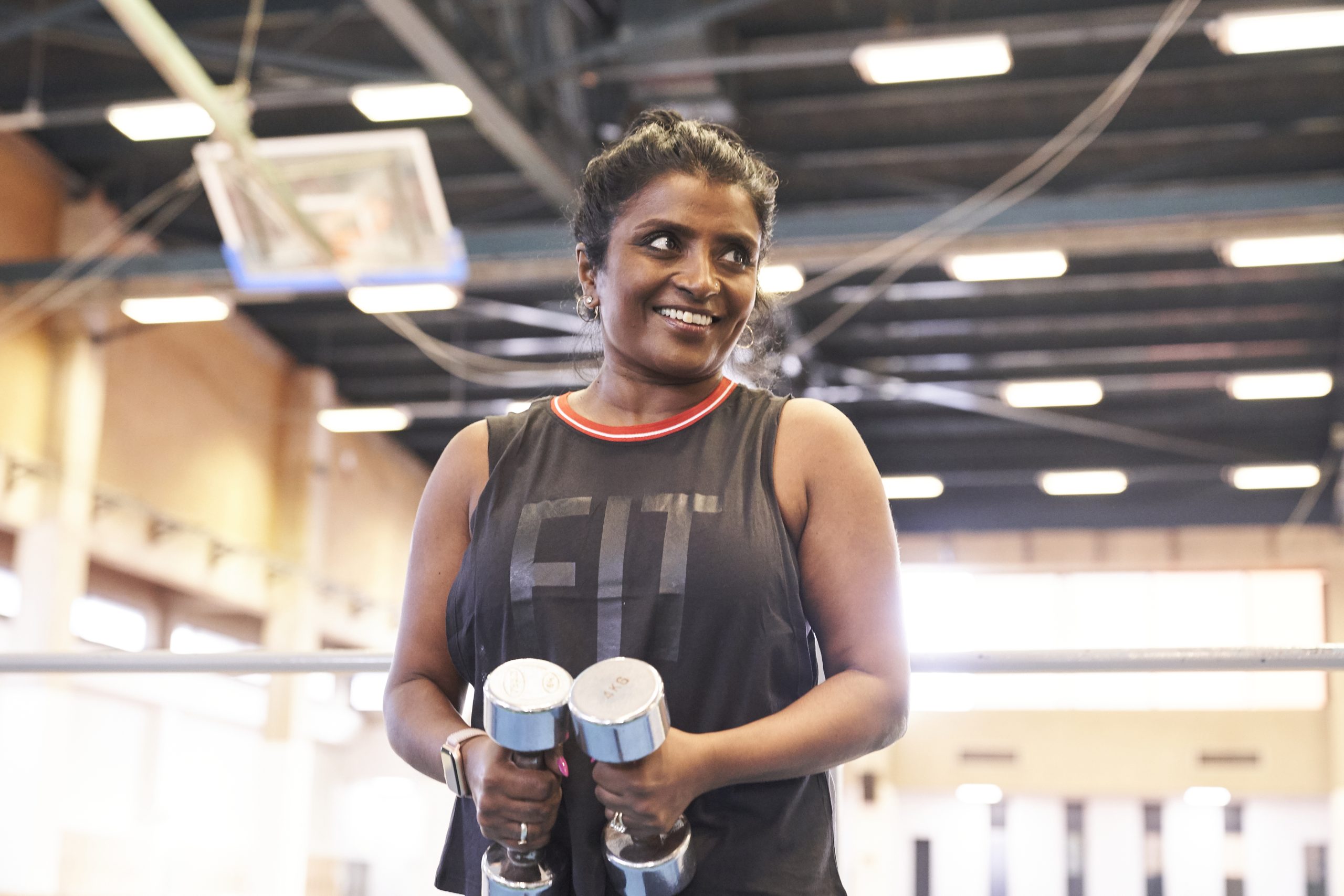 South Asian woman aged 40-50, lifting weight in a gym, smiling.