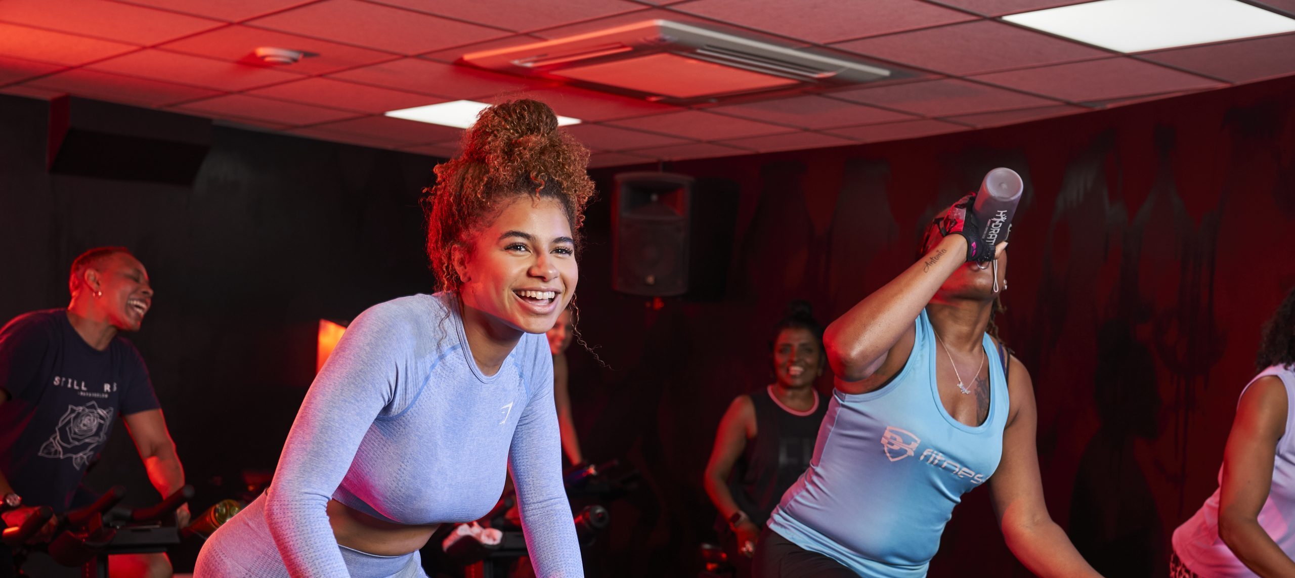 A group of women taking part in a spin cycling class. The two women at the front of the photo are Mixed Race and Black, smiling
