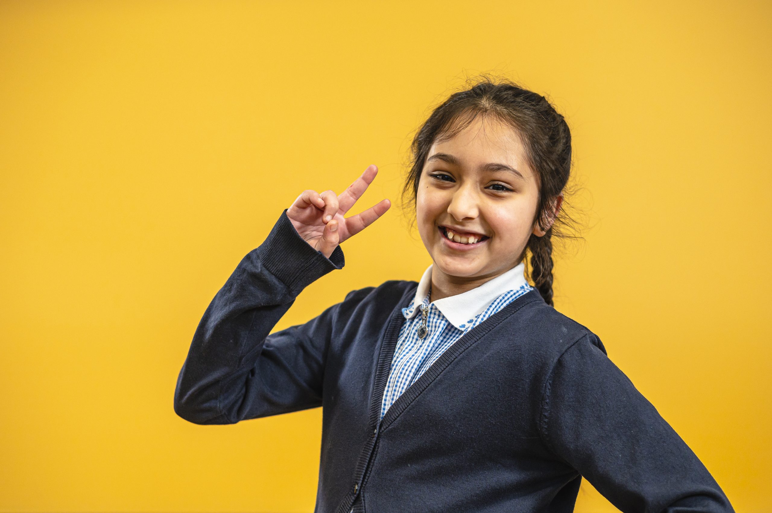 A young British south Asian girl, in her school uniform, smiling and making a peace sign